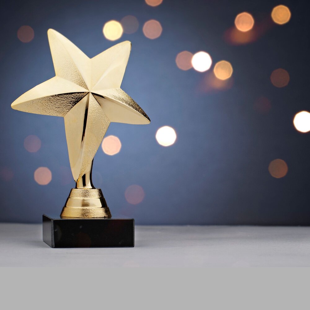 Gold star-shaped award trophy on a black base, displayed on a white surface with a blurred dark blue background and warm glowing lights. The image symbolizes achievement, recognition, and excellence.