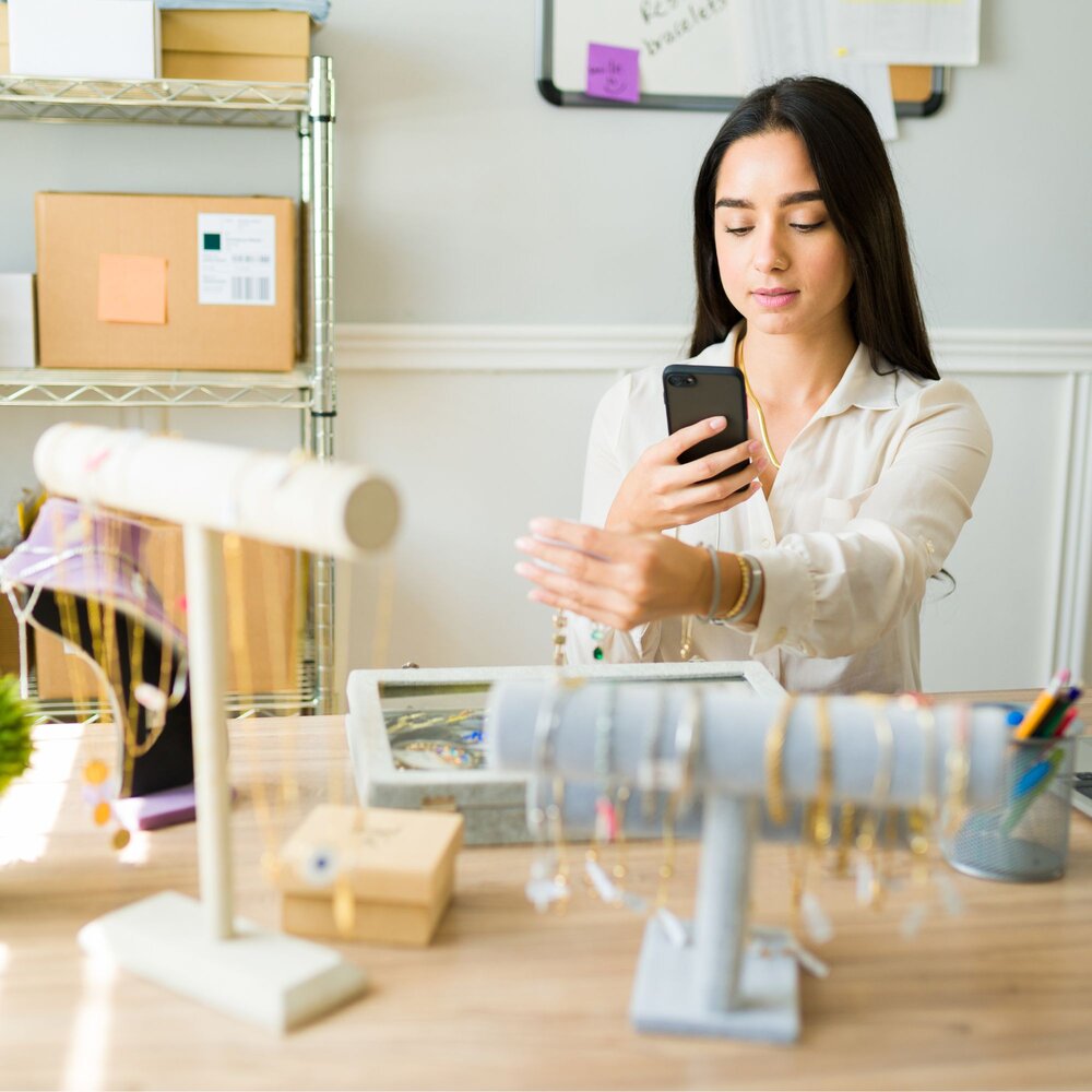 Small business owner uses a smartphone to photograph jewellery displayed on a worktable in a home or studio workspace. Jewellery stands, packaged products and storage shelves are visible around the room, illustrating product marketing, online selling and e-commerce activity within a small business setting.