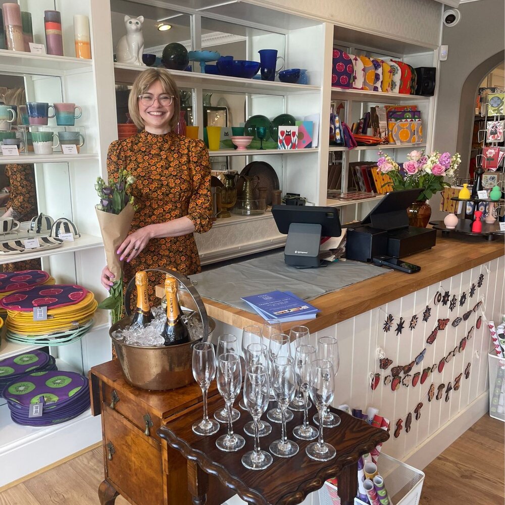 Interior of a boutique shop with colourful homeware and gifts displayed on shelves, a wooden counter with a cash register, fresh flowers in a vase, and a small table set with champagne bottles in an ice bucket and multiple empty glasses