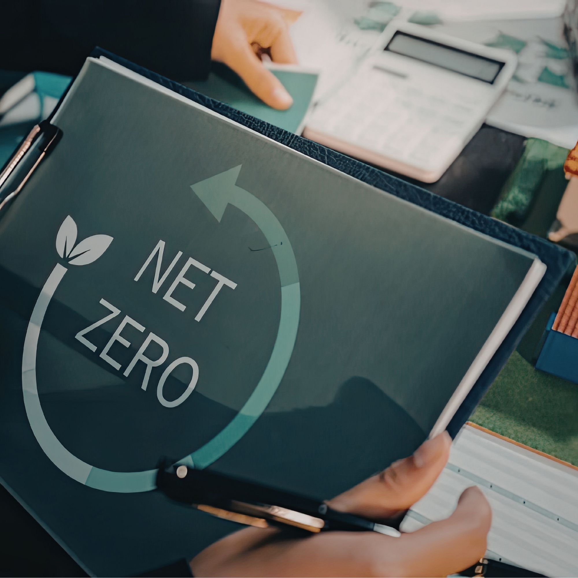 Close-up of a folder or document labelled “Net Zero,” held over a desk with stationery and papers, illustrating environmental sustainability or carbon reduction planning.