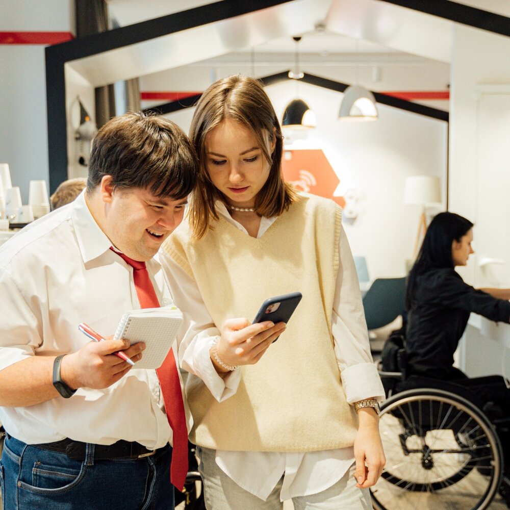 Photo of three people, man with red tie looking at a phone that a girl is holding, girl with wheelchair in the background. Office environment