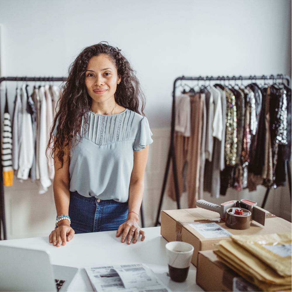 Woman wearing grey top and blue jeans stands at a shop counter with a rail of clothes behind her