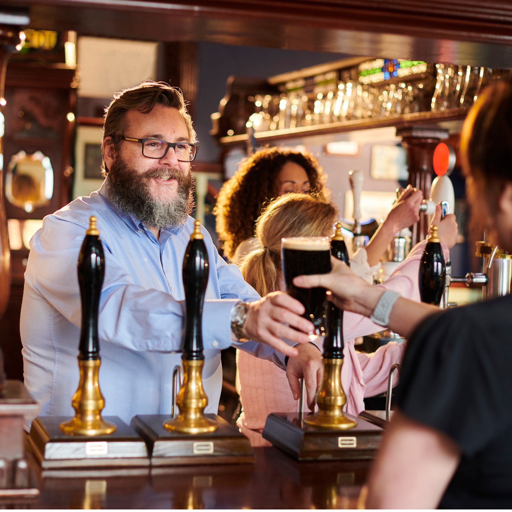 Customer receives a pint of dark beer across the bar in a traditional pub. Several people are gathered around the counter, with beer pumps and glassware visible in the foreground and background. Warm lighting and wooden furnishings create a welcoming hospitality setting.