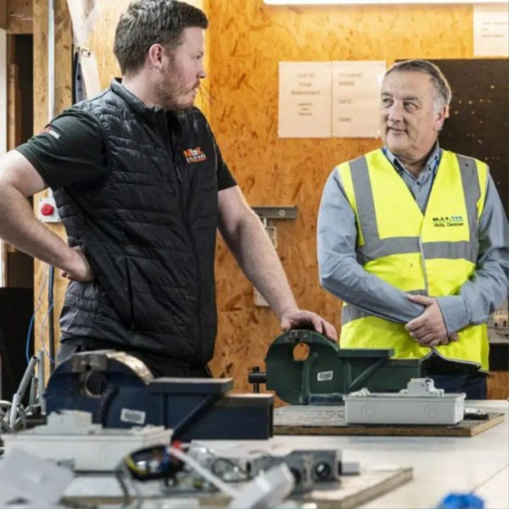 Man in black tshirt and black gilet talks to man in great shirt and high vis yellow vest