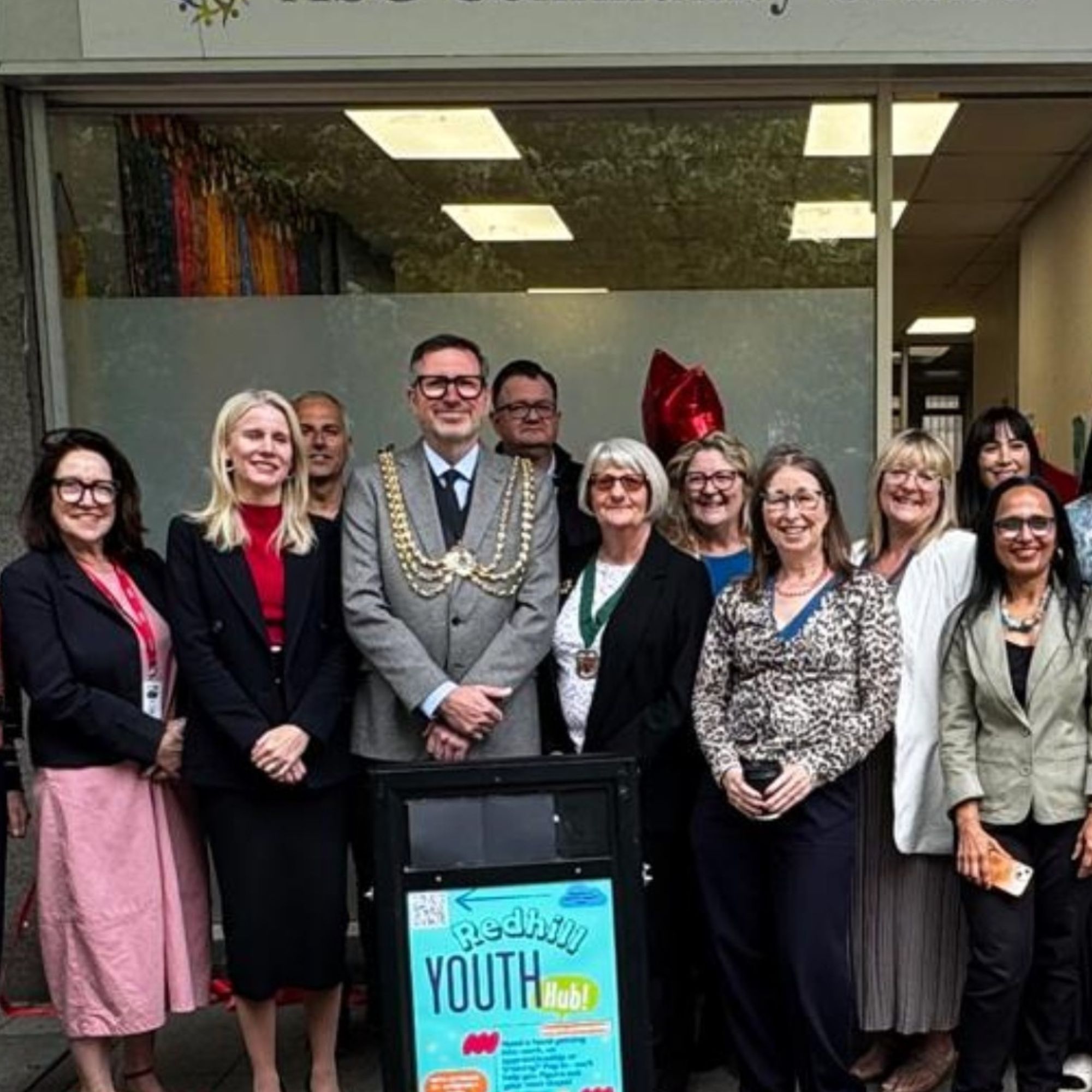 A group of adults standing outside the ASG Community Centre entrance, arranged around a sign promoting the Redhill Youth Hub. The building sign is visible above the doorway, and the group is posed on the pavement in front of the centre during a daytime event.