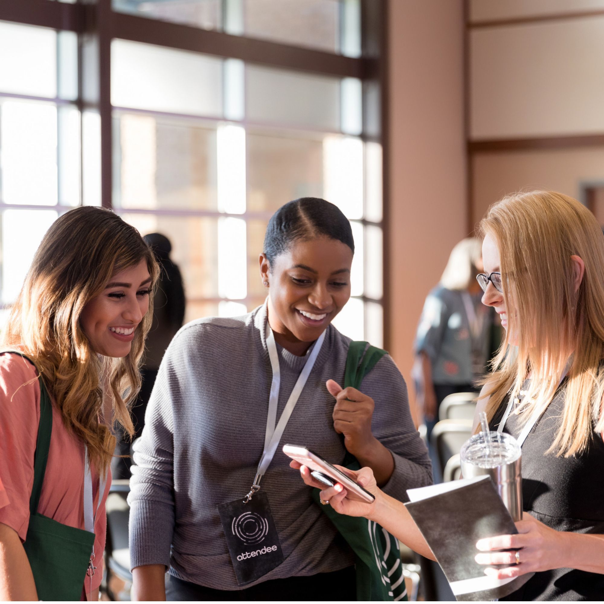 Three event attendees stand together in a bright conference or networking venue, looking at a smartphone. All three wear attendee lanyards, while one person holds a notebook and reusable drinks cup. Large windows fill the space with natural light, and additional attendees and seating are visible in the background, suggesting a business event, workshop or networking session.