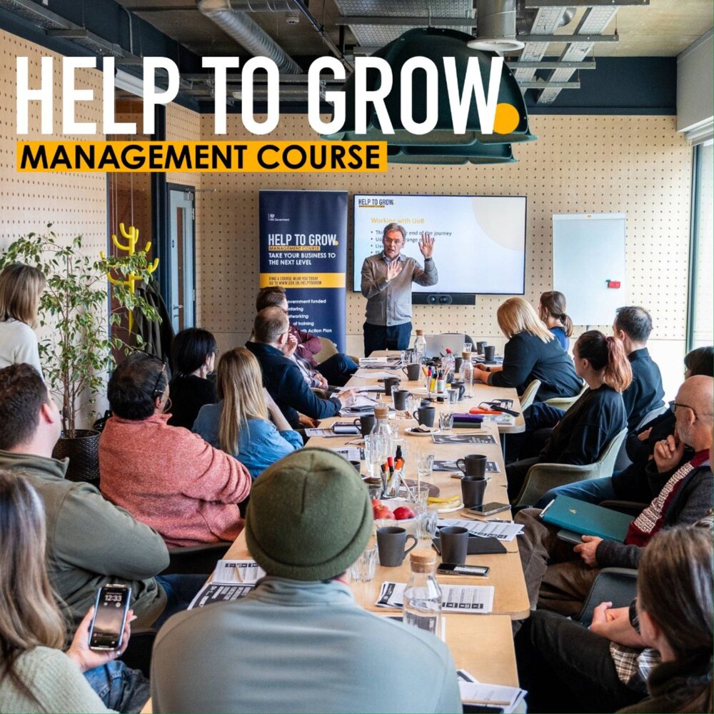 A business trainer stands at the front of a conference room presenting to a group of people seated around a long table. A large screen behind him displays a 'Help to Grow' presentation, and a banner promoting the management course is placed nearby. Attendees are listening, taking notes, and engaging with the session