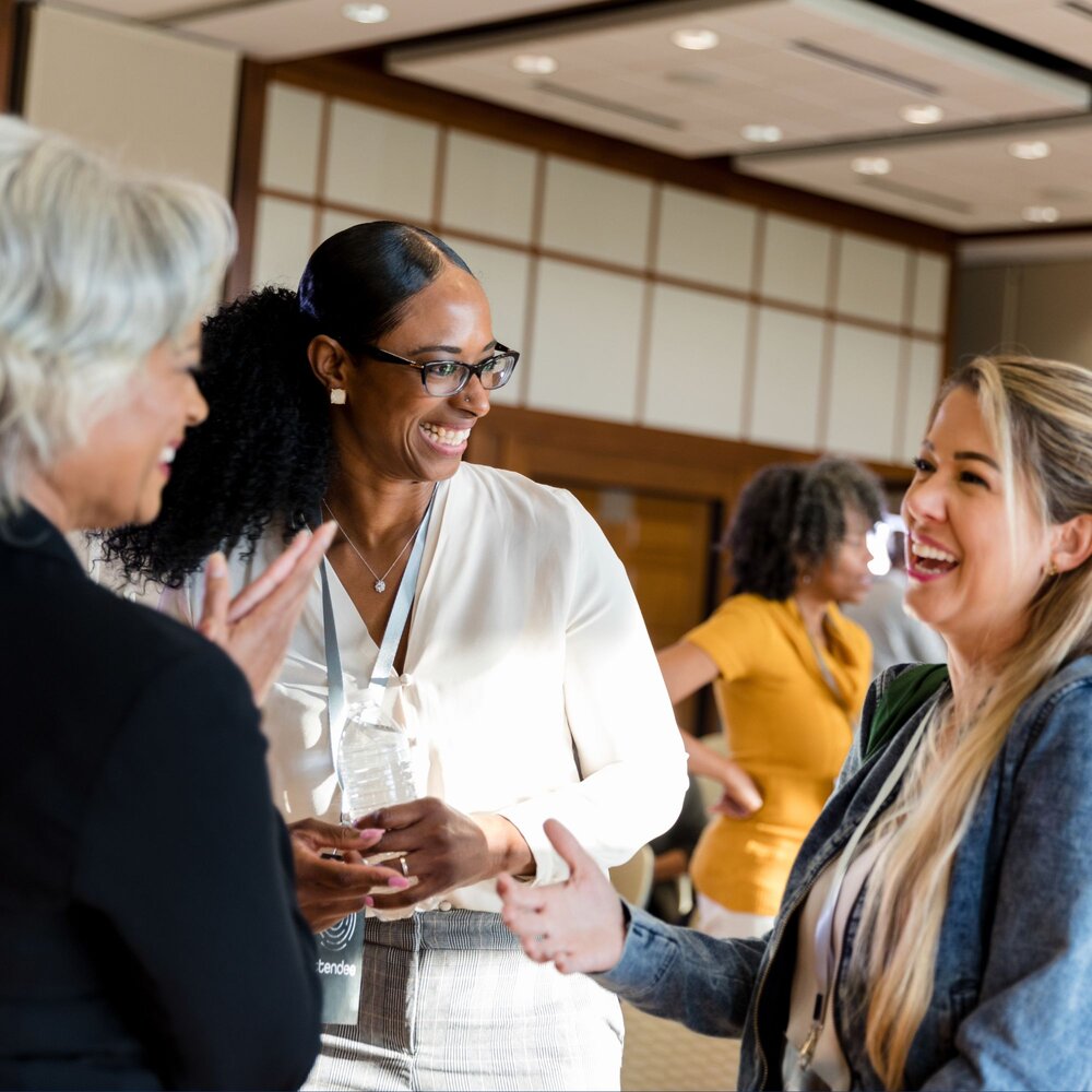 Three people wearing conference lanyards are engaged in conversation at an indoor networking event. One person in the centre, wearing glasses and a white blouse, holds a water bottle while speaking with two others in the foreground. Additional attendees are visible in the background of a well-lit meeting or conference venue.