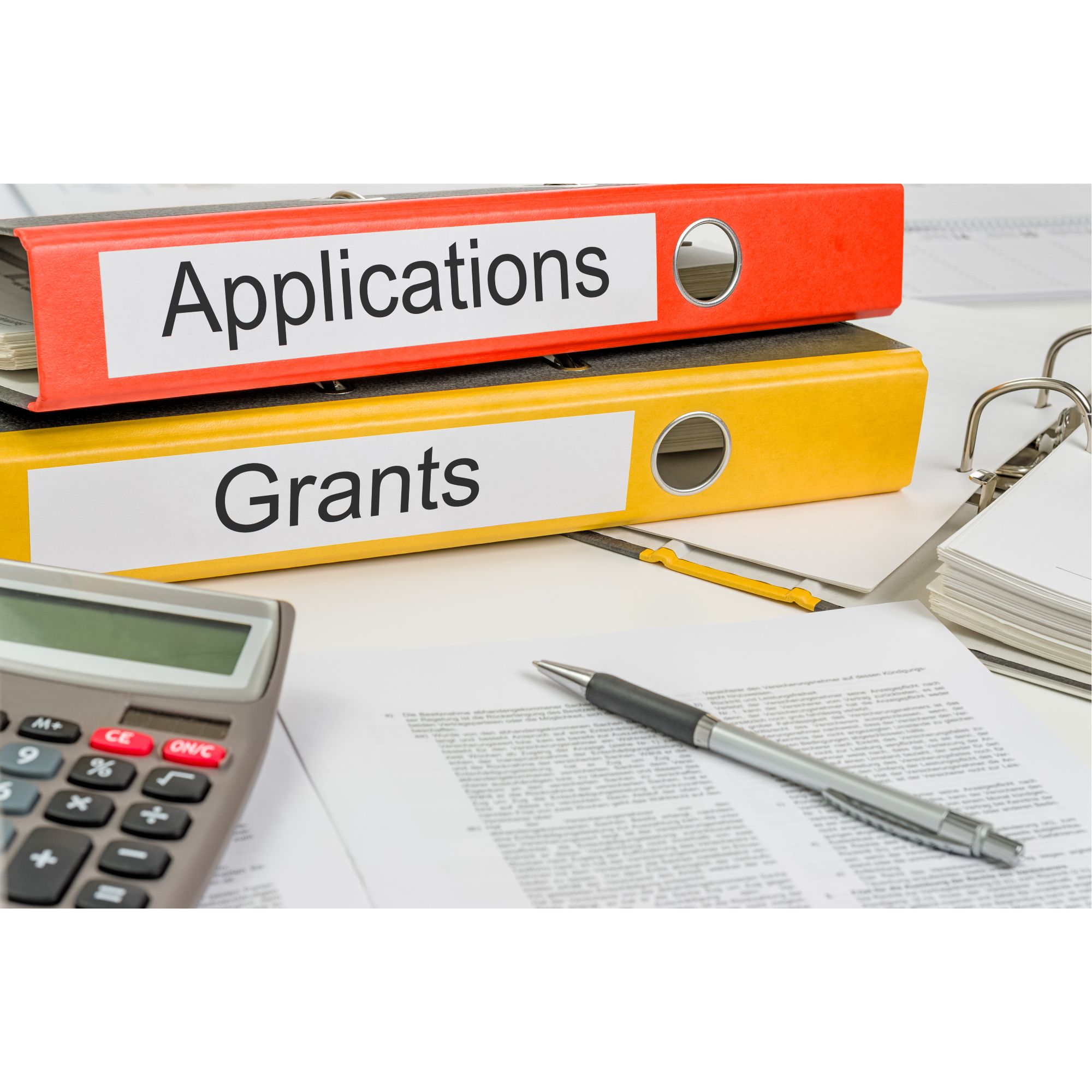Two ring binders labelled “Applications” and “Grants” stacked on a desk, surrounded by paperwork, a calculator, and a pen.