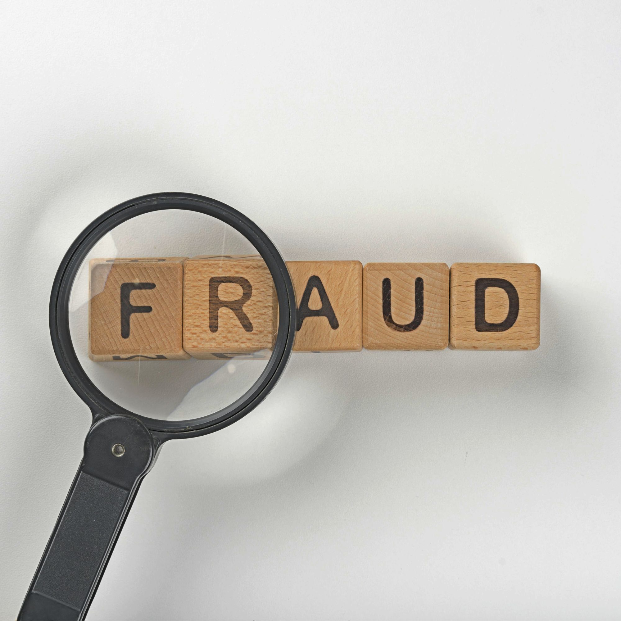 A magnifying glass positioned over wooden letter blocks spelling the word “FRAUD” on a light grey background. The magnifying lens enlarges part of the word, highlighting the letters at the beginning, creating a visual representation of fraud detection, investigation, or scam awareness.