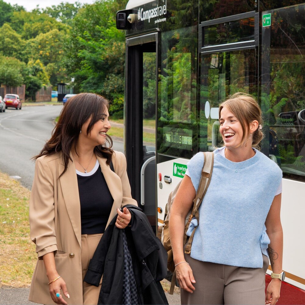 Two women walk together beside a parked bus on a quiet road lined with greenery. One wears a tan suit and carries a coat, while the other wears a light blue sleeveless top and carries a backpack over one shoulder. They appear to be in conversation as they walk away from the bus.