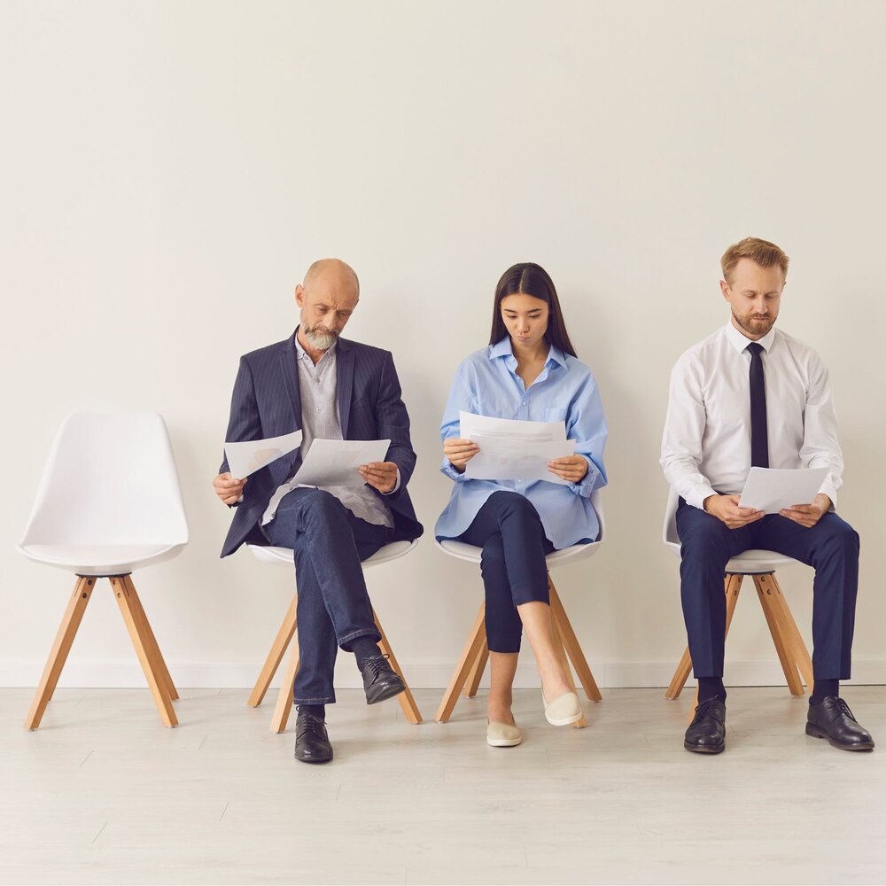 Three individuals sit on chairs against a white wall, each reviewing documents.
