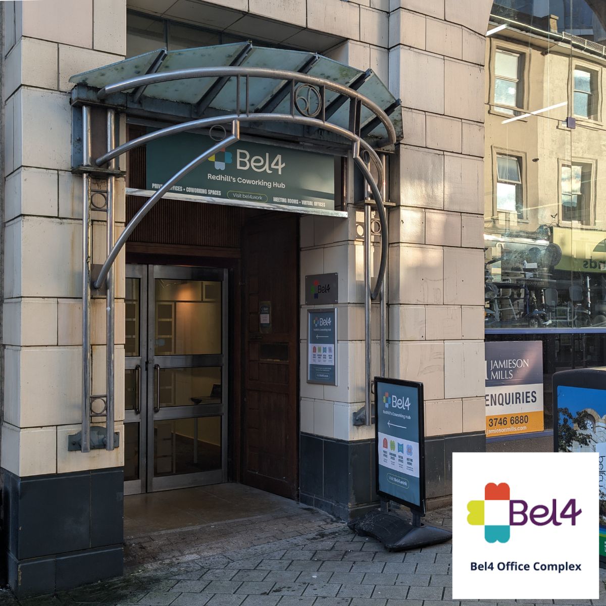 Entrance to a coworking hub named Bel4, located in a stone-fronted building with a metal canopy structure above the doorway. A glass door leads into the interior, and signage beside and in front of the entrance displays the Bel4 branding and information. Reflections of nearby buildings and shopfronts are visible in the window to the right. At the bottom-right corner of the image is the Bel4 Office Complex logo.”
