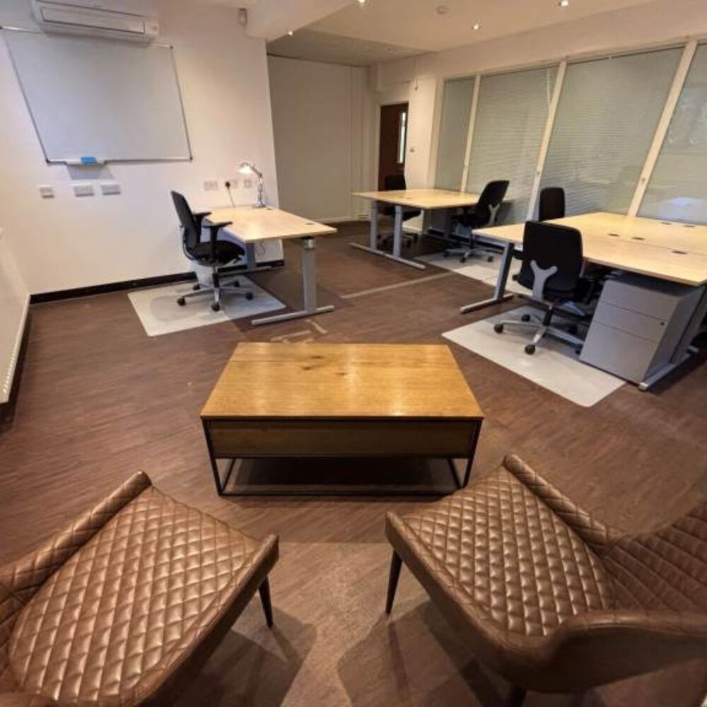 Modern office space with four desks and black office chairs arranged along the walls, each with a light wood surface and grey floor mats. In the foreground, two brown quilted chairs face a rectangular wooden coffee table with a black metal frame. The room has large windows with blinds, a whiteboard on the wall, and wood-effect flooring.”