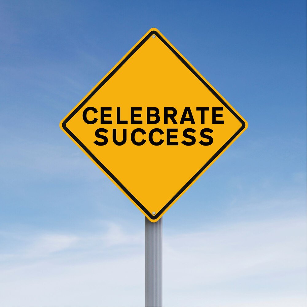 A yellow diamond-shaped road sign with the words “CELEBRATE SUCCESS” in bold black letters, set against a blue sky with light clouds.