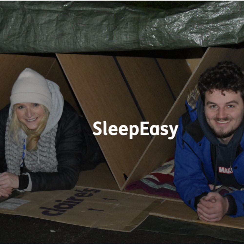 Two people lying in cardboard shelters under a tarpaulin during a SleepEasy outdoor charity event