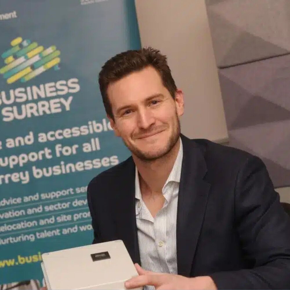 Person in business attire holding a small white box, seated in front of a Business Surrey banner offering support for Surrey businesses