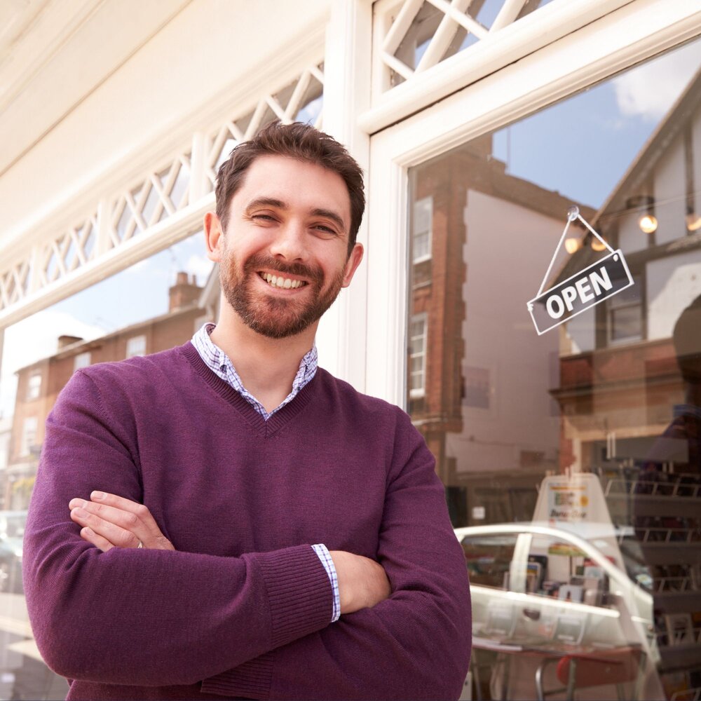 Person standing outside a high street shop with arms crossed next to an “Open” sign displayed in the window. The shopfront and neighbouring buildings are reflected in the glass, highlighting an active local business setting.