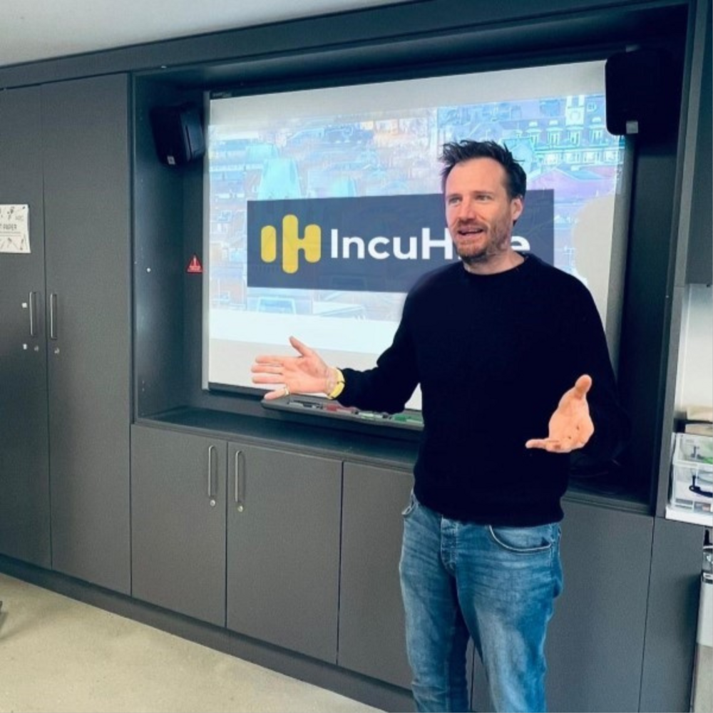 Man wearing a black jumper and jeans presenting with a screen behind him