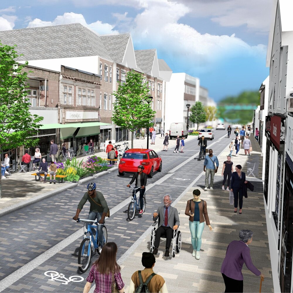 A lively urban street scene with pedestrians, cyclists, and vehicles sharing the road. The area features shops, cafes with outdoor seating, trees, flower beds, and a designated bike lane. People of varying mobility, including a wheelchair user and someone with a cane, are present, highlighting accessibility and inclusivity