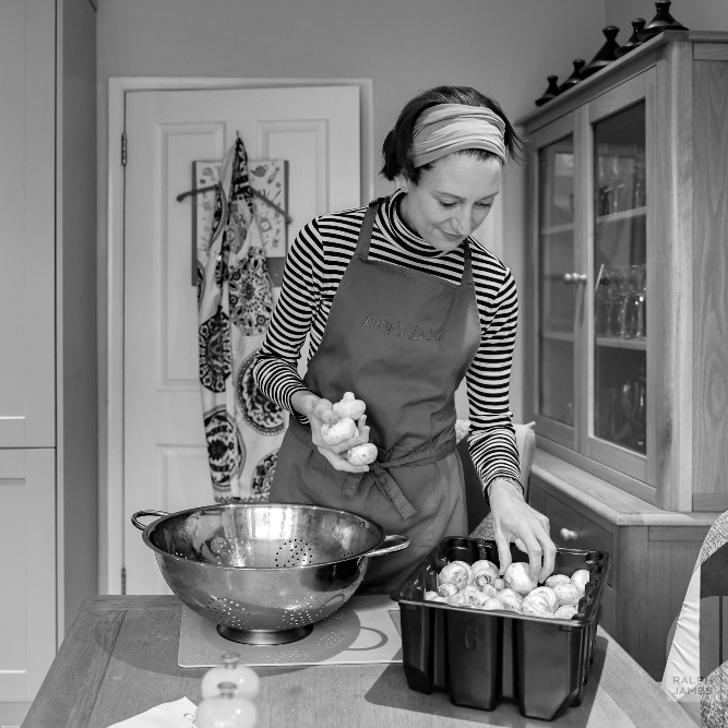 Black and white photo of woman in kitchen, with apron selecting mushrooms to wash.