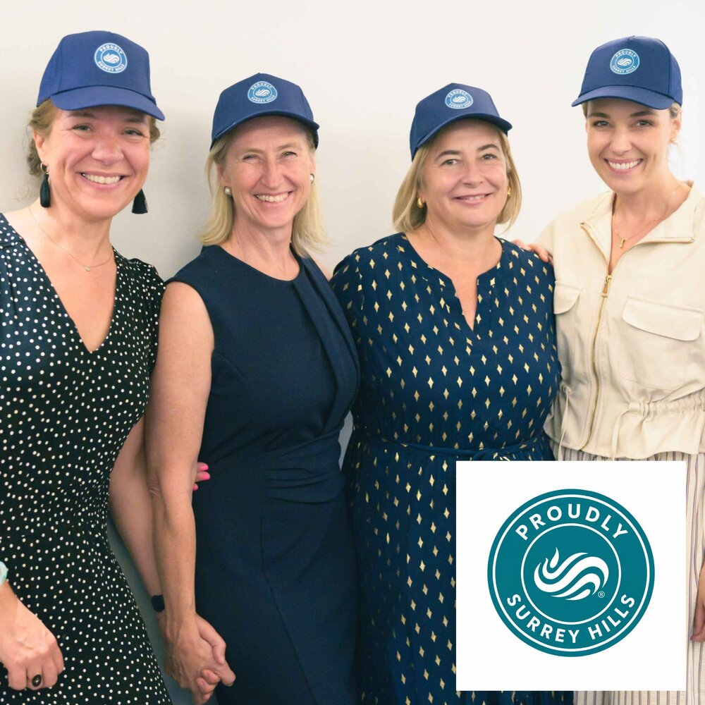 Four participants stand side by side against a plain light-coloured background, each wearing a blue cap featuring the Proudly Surrey Hills logo. The group is dressed in smart-casual attire. A large Proudly Surrey Hills logo is displayed in the bottom-right corner of the image. The photo appears to promote Surrey Hills membership, local businesses or a business networking initiative.