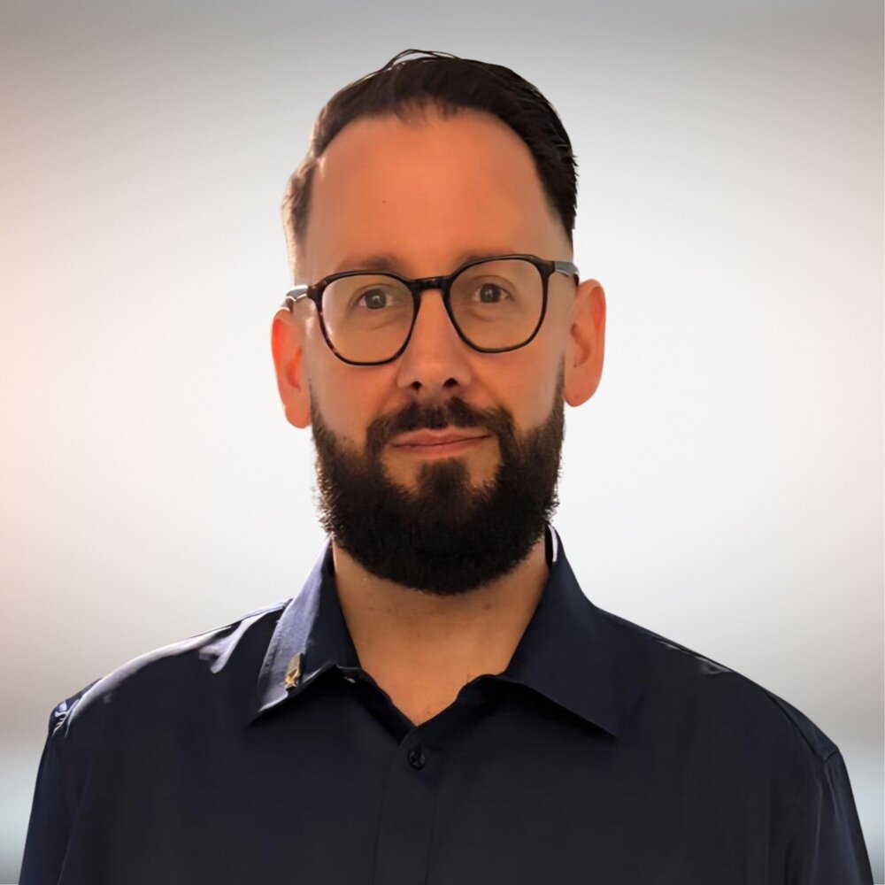 A person wearing glasses and a dark blue button-down shirt with a small, round pin.