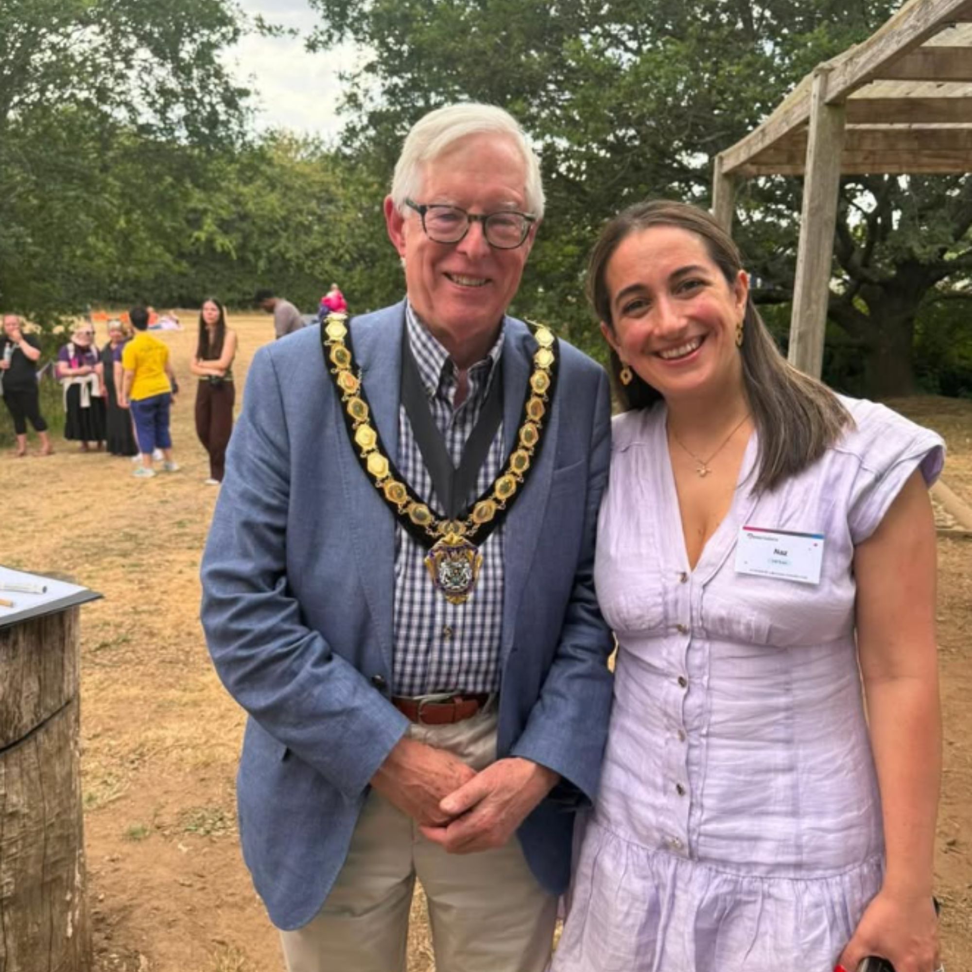 Two people stand together outdoors at a community event. One person is wearing a ceremonial chain of office over a blue blazer, checked shirt and light-coloured trousers. The other person is wearing a light purple dress with a name badge. Trees, a wooden pergola structure and several event attendees are visible in the background, with dry grass covering the ground. The photograph appears to have been taken at an informal outdoor gathering or networking event.