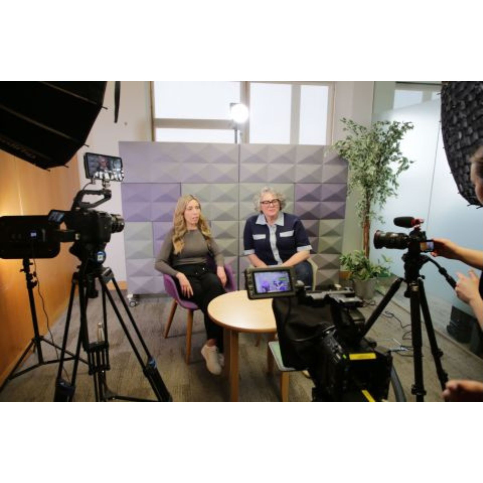 Small video interview setup with two people seated facing the camera in a studio space, surrounded by professional cameras, lights, and filming equipment, with a geometric wall backdrop and indoor plants visible.
