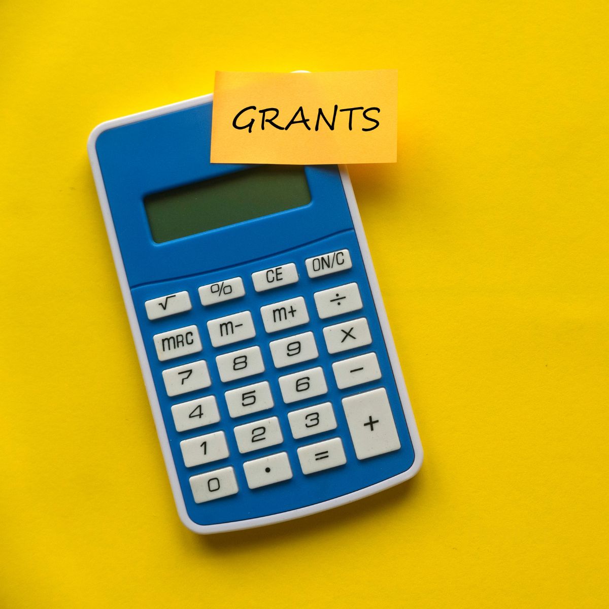 Blue calculator placed on a bright yellow background with a yellow sticky note on top that reads ‘GRANTS’ in black letters.”