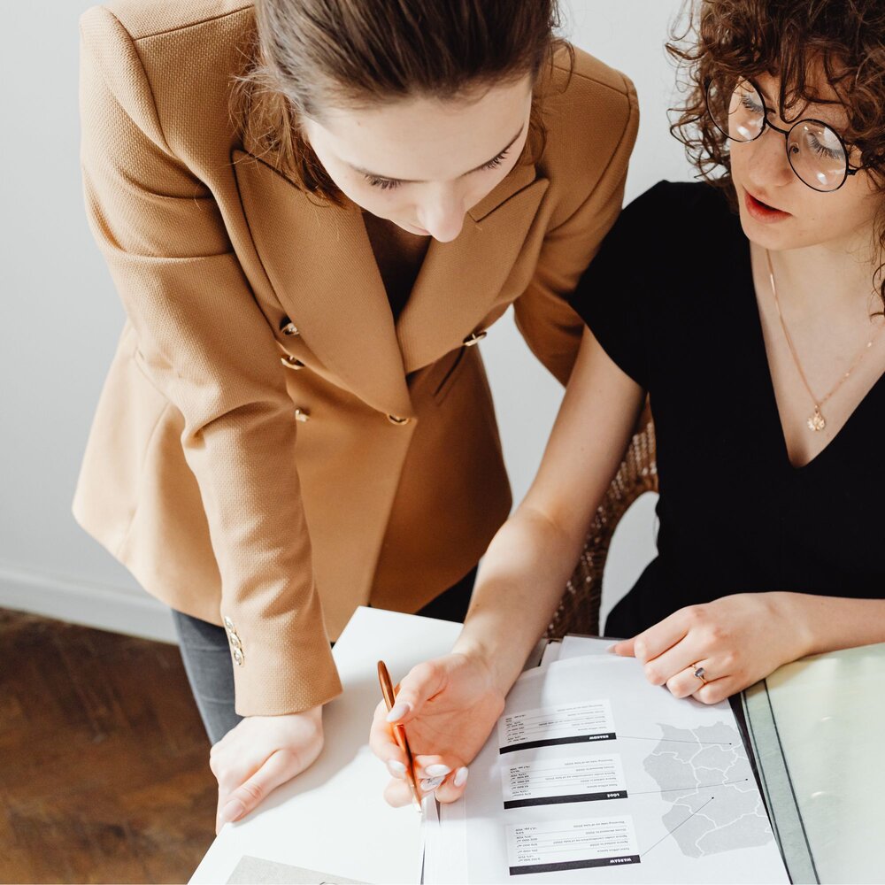 Two people collaborating at a desk, reviewing printed documents and maps, with one person holding a pen and pointing to details on the paper