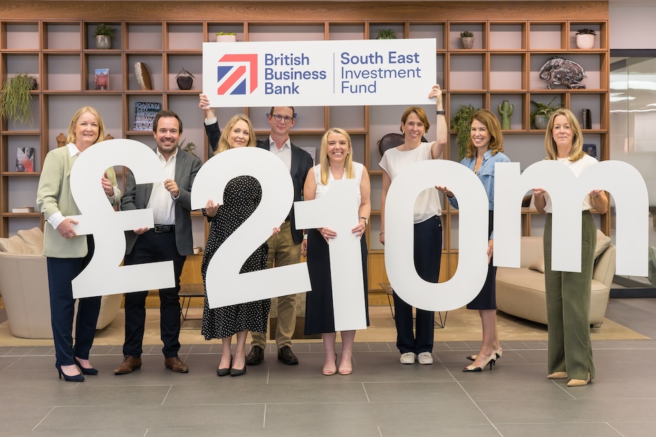 A group of people stand in an office reception area holding large white characters displaying “£210m”. Behind them, a sign reads “British Business Bank | South East Investment Fund”. Wooden shelving, seating and decorative plants are visible in the background, highlighting the launch of the £210 million investment fund for South East businesses.